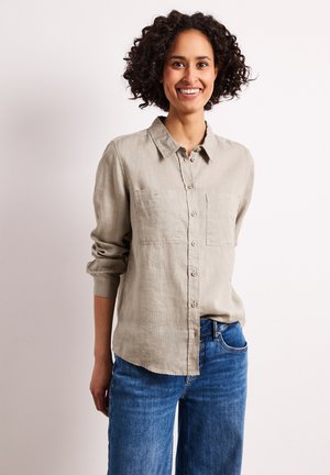 Uśmiechnięta kobieta o kręconych włosach, ubrana w beżową koszulę na guziki i niebieskie jeansy, stojąca na jednolitym jasnym tle.