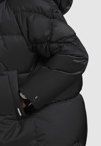 Black puffer jacket featuring a hood, quilted design, elastic cuffs, and a prominent logo detail on the sleeve. Smooth, shiny texture.