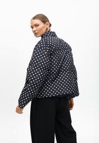 Námořnická puffer bunda s puntíky a vysokým límcem, s prošívaným designem a kontrastními světlými puntíky. Stylizováno s černými kalhotami.