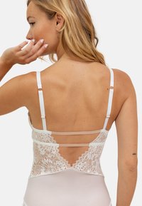 Body en dentelle avec un dos transparent, couleur nude douce, bretelles blanches réglables et un motif floral en dentelle détaillé au dos.