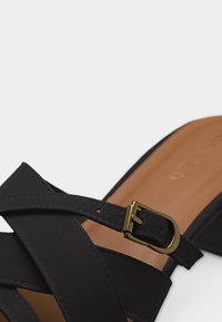 Μαύρο σανδάλι με τακούνι, με διασταυρούμενα λουράκια και χρυσή αγκράφα σε ταμπά εσωτερική σόλα, σε απλό φωτεινό φόντο.