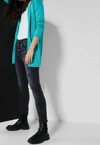 Turkos kardaig över en vit skjorta, kombinerad med svarta slim-fit jeans och svarta ankelstövlar med strukturerad yta.