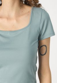 Világoskék, testhezálló felső, puha anyagból készült, négyzet alakú nyakkivágással, rövid ujjú és oldalsó hasítékokkal. Az egyik karon kerek tetoválás található.