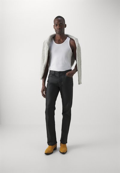Schwarze Jeans, ein weißes Tanktop und eine graue Strickjacke, die über die Schultern gelegt ist. Gelbe Wildleder-Stiefeletten setzen einen kontrastreichen Akzent.