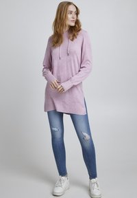 Hoodie rose clair en tissu doux, avec un cordon de serrage, des manches longues et des fentes sur les côtés. Assorti à un jean skinny bleu déchiré et des baskets blanches.