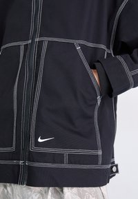 Casaco preto feito de tecido com costuras brancas. Apresenta um fecho na frente, bolsos laterais e um pequeno logótipo da Nike perto da barra.