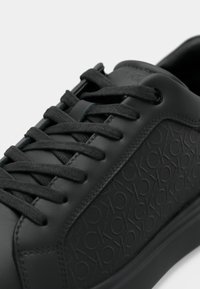 Zapatilla negra con un patrón de logo en relieve con la repetición de 'ck' en el panel lateral y cordones negros pasados por seis ojetes.