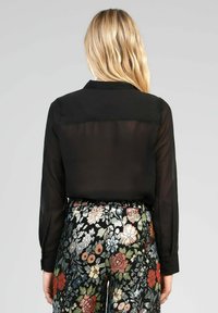 Blouse noire transparente avec des manches longues et un col, associée à un pantalon texturé à motif floral avec des fleurs colorées.