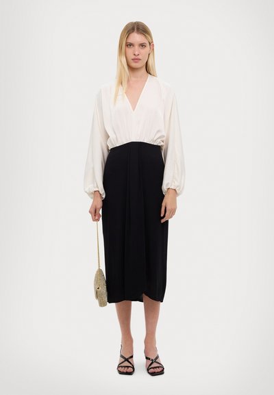 Blouse blanche à manches longues avec un décolleté en V plongeant ; jupe noire jusqu'aux genoux ; modèle tenant un petit sac à main texturé de couleur dorée ; sandales noires à lanières.