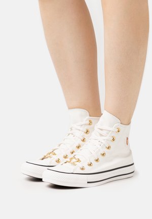 Białe sneakersy typu high-top wykonane z płótna, z złotymi oczkami, czarnymi paskami na podeszwie i haftowanym sercem po zewnętrznej stronie.