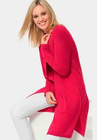 Gilet en tricot rose à ouverture sur le devant, à manches longues et avec deux poches latérales, porté par-dessus un pantalon blanc. Texture lisse et design mi-long.