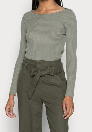 Vrouw draagt een geribd, langärmlig, saliegroene top, ingestopt in een hooggehuisde olijfgroene broek met een ceintuur die aan de taille is gestrikt.