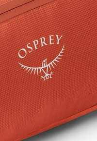 Borsa di stoffa rossa con superficie testurizzata, con un logo OSPREY bianco e un'immagine di un'aquila al centro. Cerniera lungo il bordo superiore.