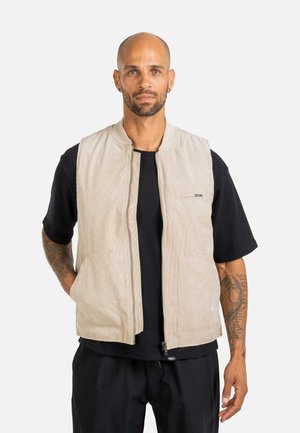 Man with tattoos wearing a beige sleeveless zip-up vest over a black t-shirt, standing with hands in pockets against a white background.