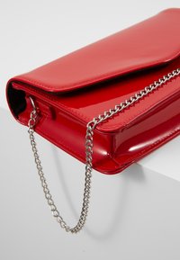 Rød håndtaske i laklæder med en fold-over klap og sølvkæde som rem. Tasken har en glat, skinnende tekstur og en struktureret form.