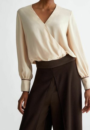 Frau trägt eine cremefarbene, langärmelige Bluse mit V-Ausschnitt, gerafften Manschetten und eine hoch taillierte, dunkelbraune, weit geschnittene Hose mit vorderen Schlitzen.