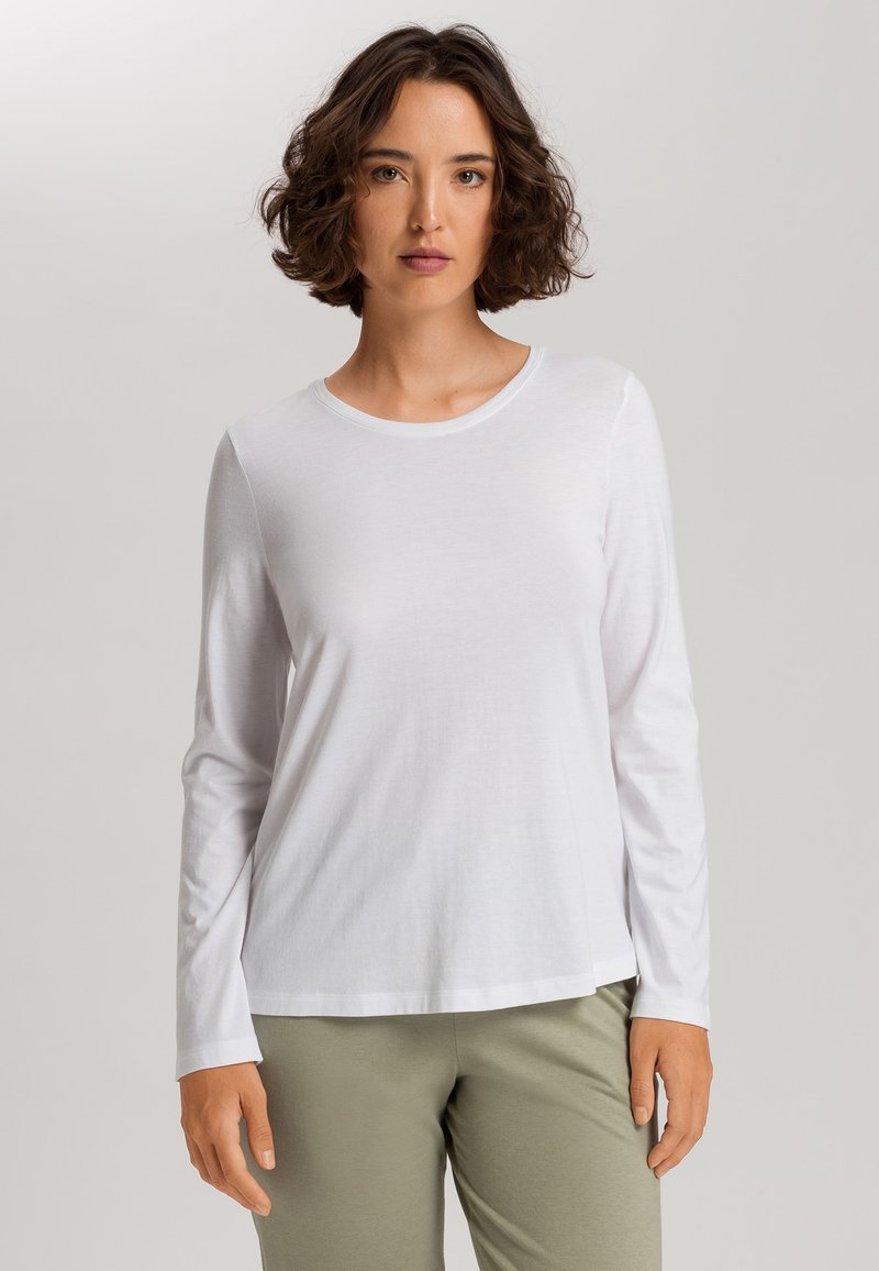 Camisa blanca de manga larga y cuello redondo, hecha de una tela suave, con un corte relajado y un dobladillo ligeramente alargado, combinada con pantalones verde claro.