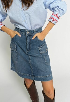 Mujer con una camisa azul a rayas con puños bordados, una falda vaquera azul con bolsillos delanteros y botas marrones de cuero hasta la rodilla.
