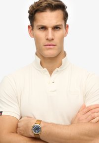 Polo shirt crema con colletto aperto, tessuto texurizzato e patch del logo; accessoriato con un orologio dorato e cinturino in cuoio beige.