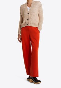 Béžový pletený cardigan se třemi černými knoflíky, kombinovaný s jasně oranžovými kalhotami širokého střihu a robustními černými dřeváky na podpatku.