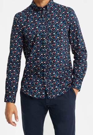 Homme portant une chemise à manches longues avec un motif géométrique abstrait bleu, rouge et blanc et un pantalon bleu foncé, main dans la poche.
