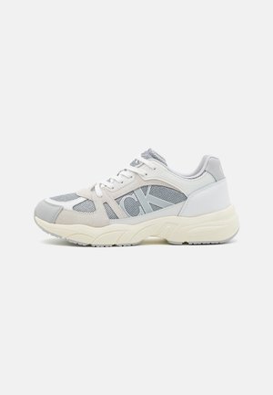 Sneaker grigie chiare con pannelli in rete e suede, caratterizzate da un logo prominente, suola testurizzata e accenti bianchi. Punta arrotondata e design imbottito.