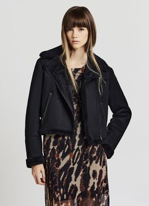 Giovane donna con capelli castani lisci che indossa una giacca in shearling nera sopra un lungo vestito con stampa animalier su uno sfondo uniforme.