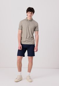 Kaki polo majica z zadrgo na ovratniku, v paru z mornarsko modrimi kratkimi hlačami. Model nosi svetle športne copate in bele nogavice, stoji pred nevtralnim ozadjem.