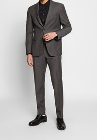 Szary garnitur z teksturowanej tkaniny, marynarka jednorzędowa z dwoma guzikami, w połączeniu z czarną koszulą i czarnymi butami.