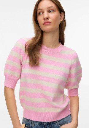 Vrouw draagt een kort shirt met roze en beige strepen en opgeblazen mouwen, in combinatie met spijkerbroeken, kijkt opzij tegen een witte achtergrond.