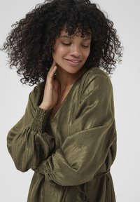 Femme aux cheveux bouclés portant une robe longue à manches longues de couleur vert olive brillant, touchant doucement son cou et souriant les yeux fermés.