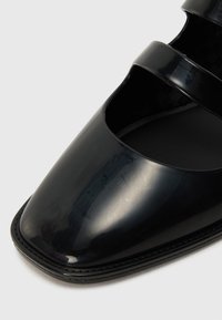 Black patent leather shoe with a glossy finish, angular shape, open cutouts on sides, and contrasting stitching along the sole.