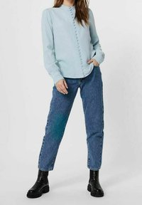 Himmelbluse mit langen Ärmeln, Stehkragen und Knopfdetails, kombiniert mit hochgeschnittenen blauen Jeans und schwarzen Stiefeletten.