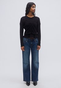 Μαύρο μακρυμάνικο τοπ με ραβδώσεις και εμπρός σχεδιασμό με κόμπο, σε συνδυασμό με τζιν μπλε τζιν παντελόνι με ψηλή μέση και φαρδιά μπατζάκια και μαύρα παπούτσια με μυτερή μύτη.