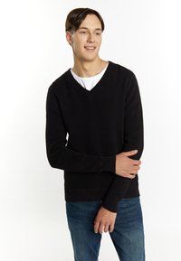 Pulover negru, texturat, cu decolteu în V și mâneci lungi, asortat cu blugi albaștri. Materialul are un design canelat și manșete adaptate.