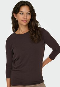 Maglietta marrone a maniche lunghe con scollo rotondo e vestibilità comoda. Realizzata in tessuto morbido con una superficie liscia. Il modello è posato su uno sfondo neutro.