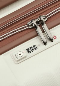 Valigia rigida marrone con zip a doppio tirante metallico, sfondo in tessuto bianco e un lucchetto a combinazione montato sul lato con un foro per la chiave.