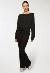 Felpa nera oversize con colletto scoperto, abbinata a pantaloni neri svasati e sneakers di colore chiaro. Texture liscia.