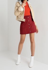 Giacca di velluto a coste beige con colletto in shearling bianco, top rosso aderente, gonna bordeaux con bottoni e stivali bianchi alla caviglia.