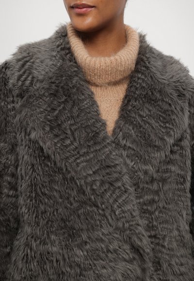 Pilkos dirbtinės vilnos paltas su tekstūruota paviršiaus struktūra, turintis platų apykaklę. Dėvimas ant šviesiai smėlinio trikotažo turtleneko megztinio.