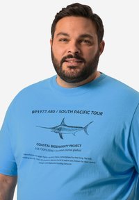 Camiseta de algodón de color azul claro con una ilustración de pez espada y texto que detalla el Proyecto de Biodiversidad Costera. Diseño sencillo de cuello redondo.