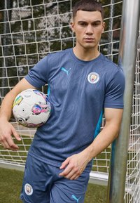 Zils sporta krekls ar teksturētu rakstu un tirkīza akcentiem, kopā ar saskaņotām šortiem. Spēlētājs tur rokās krāsainu, rakstainu futbola bumbu.