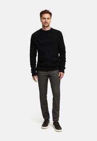 Czarny sweter w splotrze warkoczowym, szare spodnie o teksturowanym wykończeniu oraz ciemne sneakersy. Model stoi na białym tle, ręce swobodnie wzdłuż ciała.