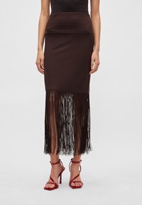 Bruine, nauwsluitende rok met een gladde bovenkant en lange, vallende franjes aan de zoom. Gecombineerd met rode sandalen met hoge hak en bandjes.