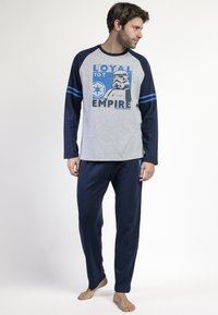 Hombre vistiendo un conjunto de pijama de manga larga en navy y gris de Star Wars con la frase "Leal al Imperio" y un gráfico de Stormtrooper, de pie descalzo mirando hacia la derecha.