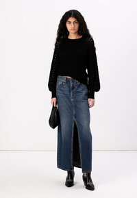 Maglione nero lavorato a maglia con maniche strutturate, abbinato a una gonna maxi in denim blu a vita alta con uno spacco frontale. Borsa nera e stivaletti alla caviglia.