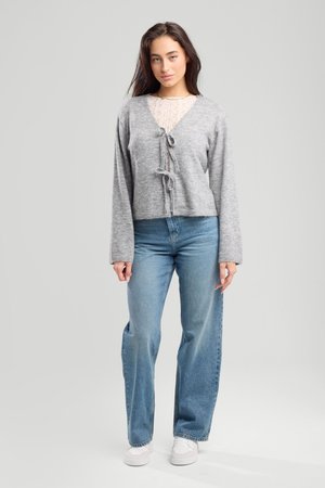 Giovane donna con lunghi capelli scuri che indossa un cardigan grigio annodato davanti, top in pizzo, jeans blu e sneakers bianche, in piedi davanti a uno sfondo neutro.