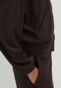 Dark brown athletic jacket featuring a soft texture, elastic cuffs, a side pocket, and a relaxed fit. Tailored design with minimal detailing.