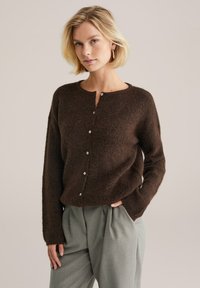 Cardigan maro, texturat, cu guler rotund și mâneci lungi, având o serie de nasturi albi pe față. Asortat cu pantaloni gri deschis.