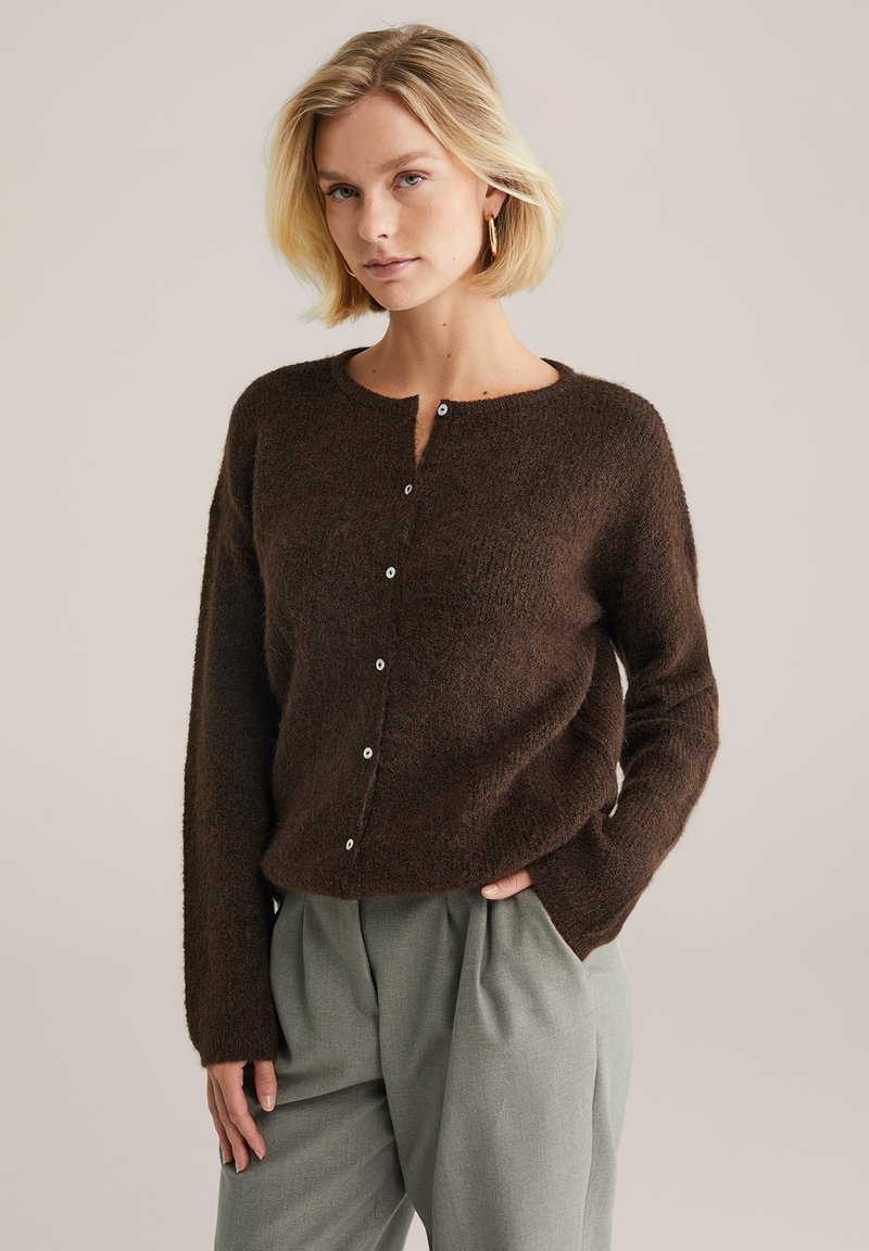 Cardigan maro, texturat, cu guler rotund și mâneci lungi, având o serie de nasturi albi pe față. Asortat cu pantaloni gri deschis.